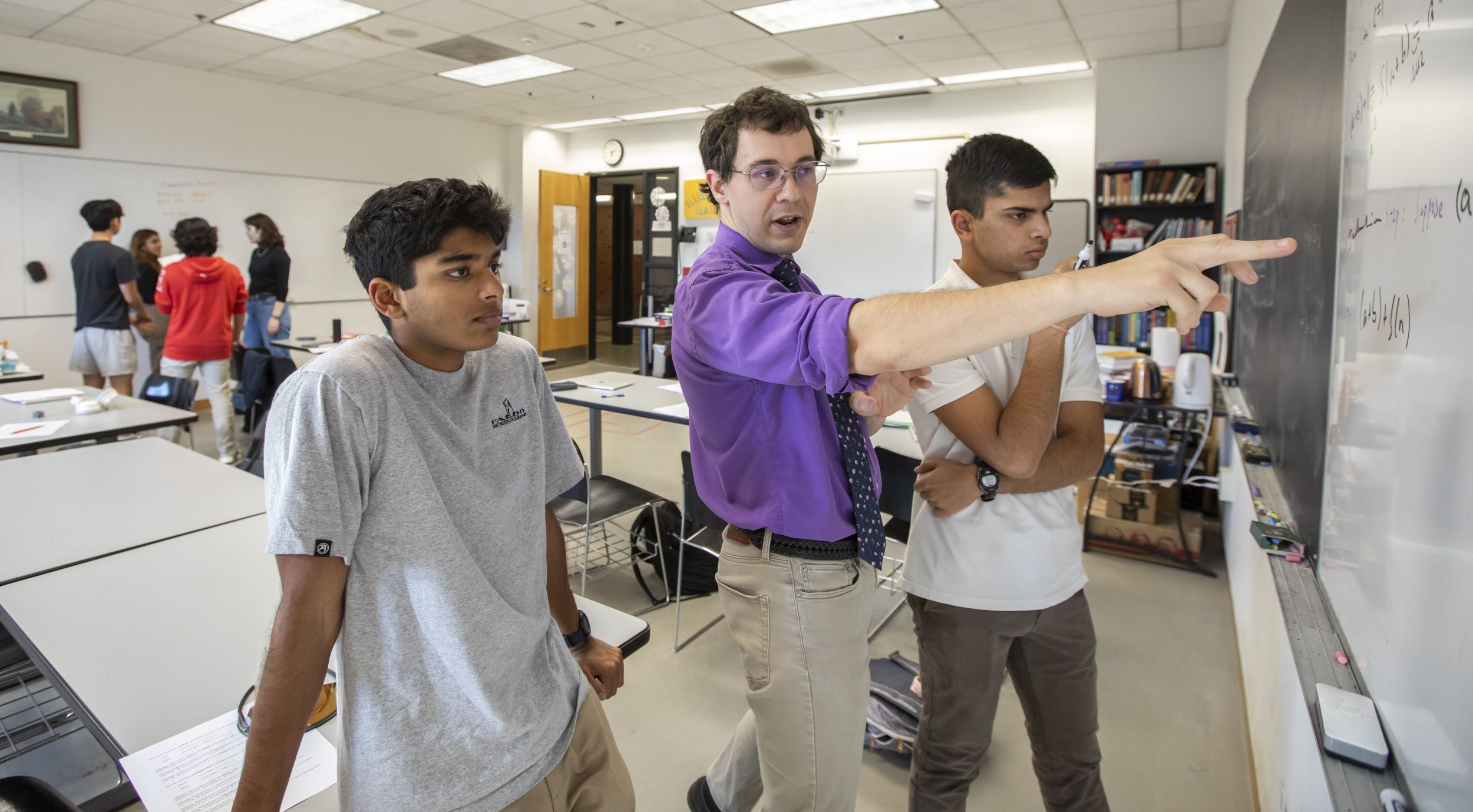 Math teacher Anton Fleissner, wearing a purple dress short with the sleeves rolled up, points to an equation on the chalkboard while two students look on.
