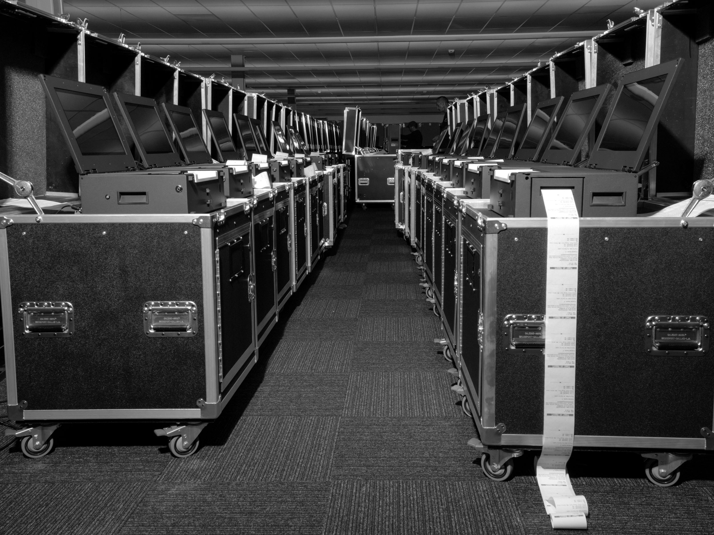 Black and white photo of two rows of Clar Ballot voting machine scanners, with a white paper tape of logged activity unspooling from the one in the front right.