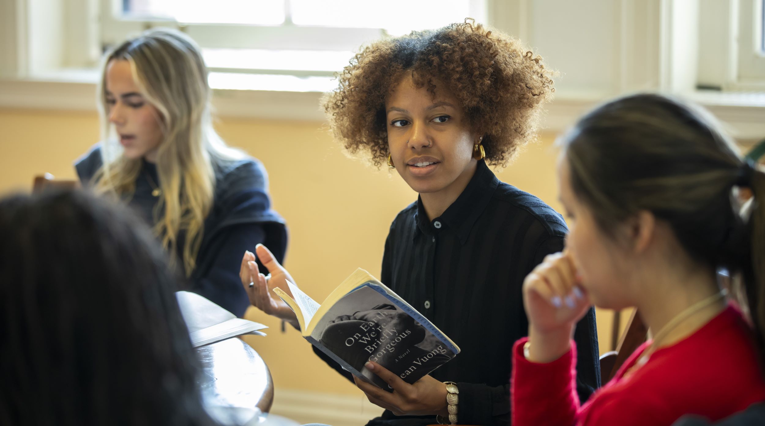English teacher Victoria Still holds a paperback novel while holding her upturned right hand out to help illustrate a point and her students at the Harkness table listen.