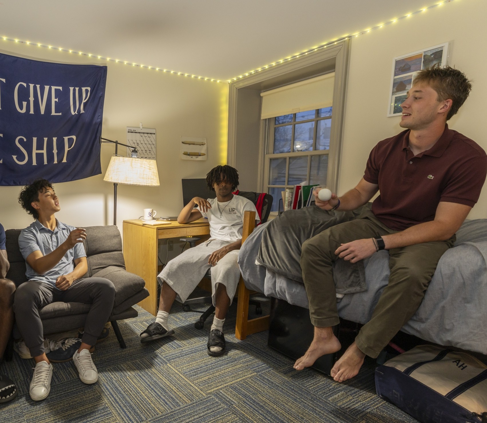 An older student sits on the top of a bunk bed in a dorm room, speaking, while three younger students pay attention to him.