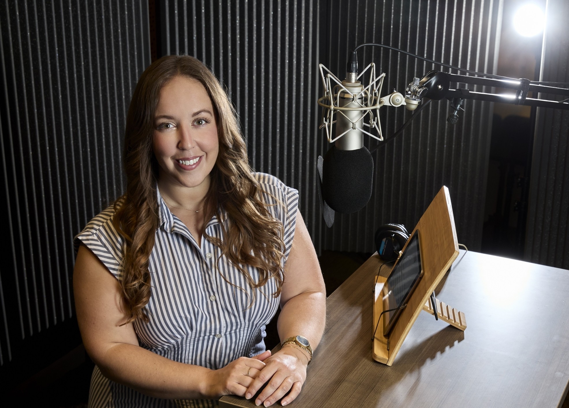 A woman with long brown hair stands before a hanging microphone in a recording studio. She is smiling with her hands placed on a brown wood-grain table.