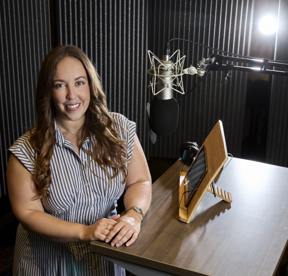 A woman with long brown hair stands before a hanging microphone in a recording studio. She is smiling with her hands placed on a brown wood-grain table.