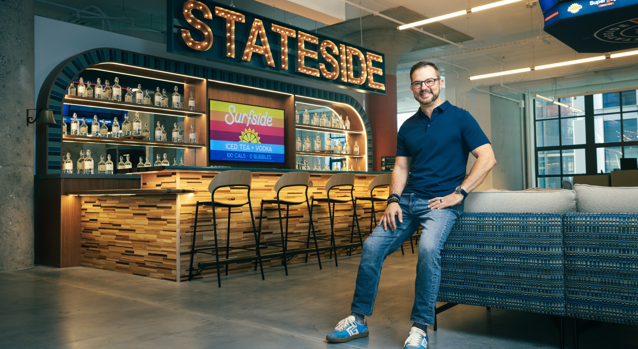 Clem Pappas, in a navy blue polo shirt and jeans, sits on the back of a blue couch with his hands resting on his upper thighs inside the bar area of his Stateside Brands headquarters. There is a long bar with a row of empty seats behind him, and in block letters formed by clear light bulbs, it spells out "STATESIDE" above the bartenders' area.