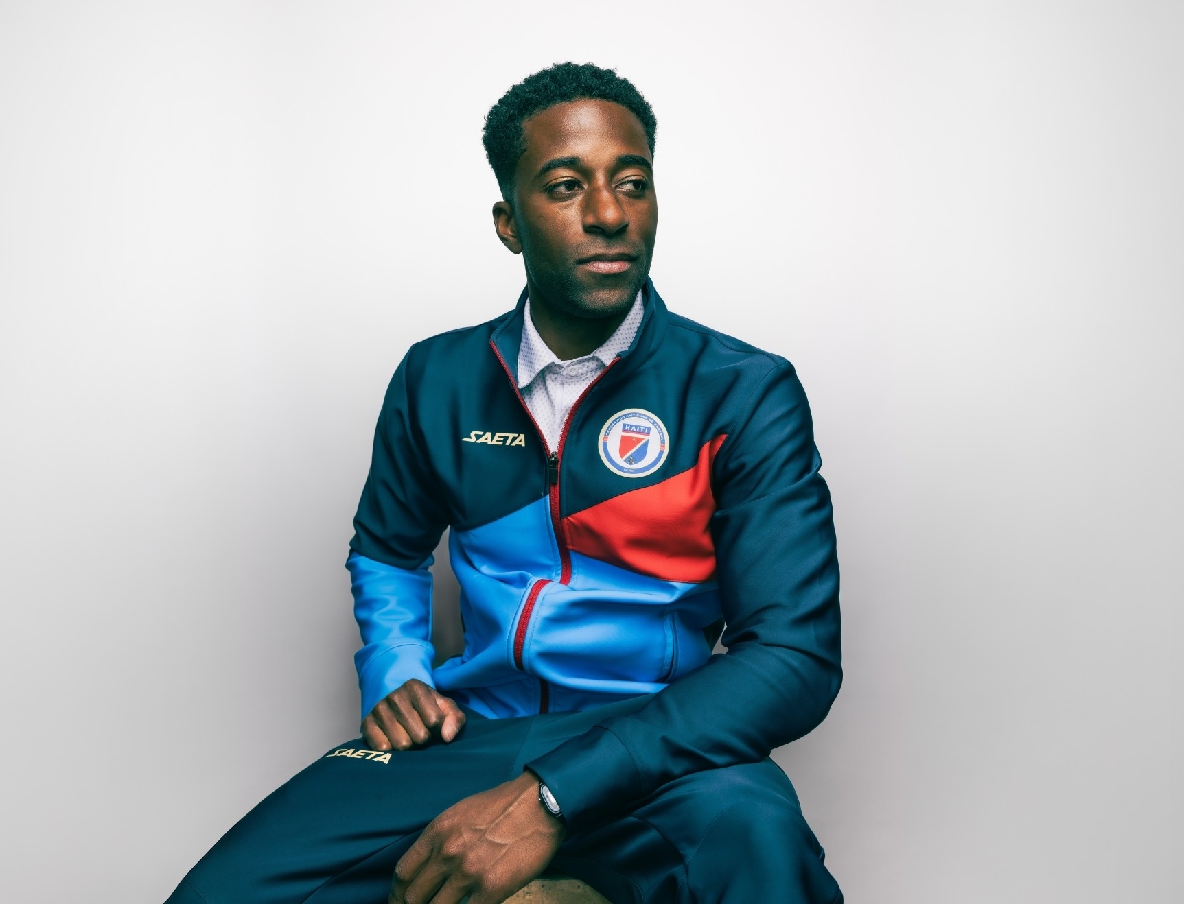 Duke Lacroix, wearing a dark blue, light blue, and red sweatsuit, sits on a stool with his hands resting in front of him. He is expressionless and looking slightly downward. The background is plain white.