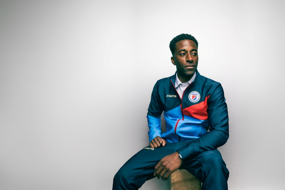 Duke Lacroix, wearing a dark blue, light blue, and red sweatsuit, sits on a stool with his hands resting in front of him. He is expressionless and looking slightly downward. The background is plain white.