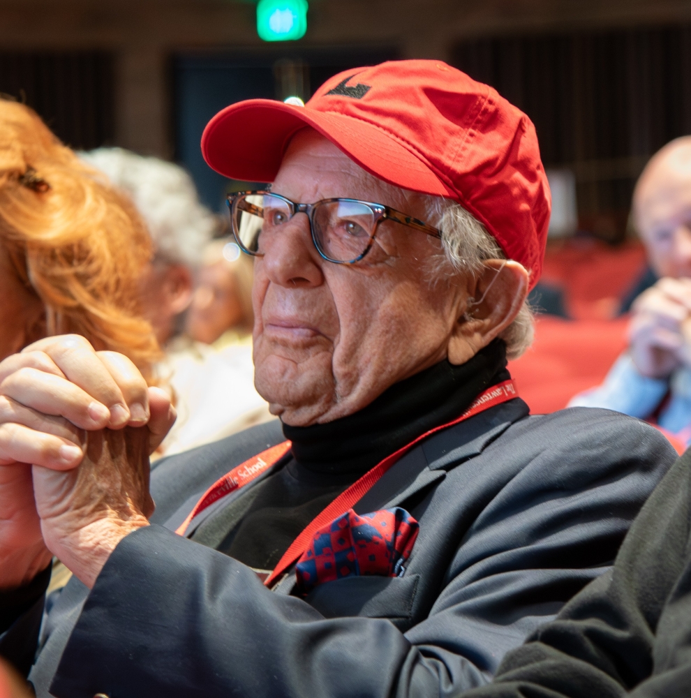 An older man wearing a red cap with a black L on it, glasses, a dark turtleneck and a dark blazer, clasps his hands while seated and looking up at something.